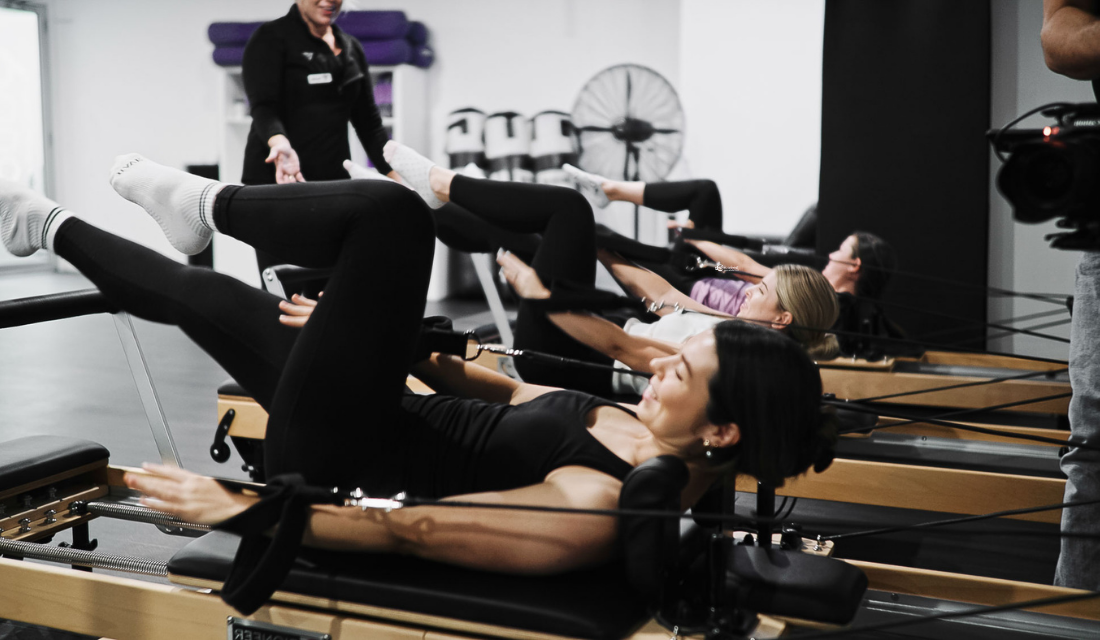 An image of three women using pilates reformers while being instructed by a teacher.