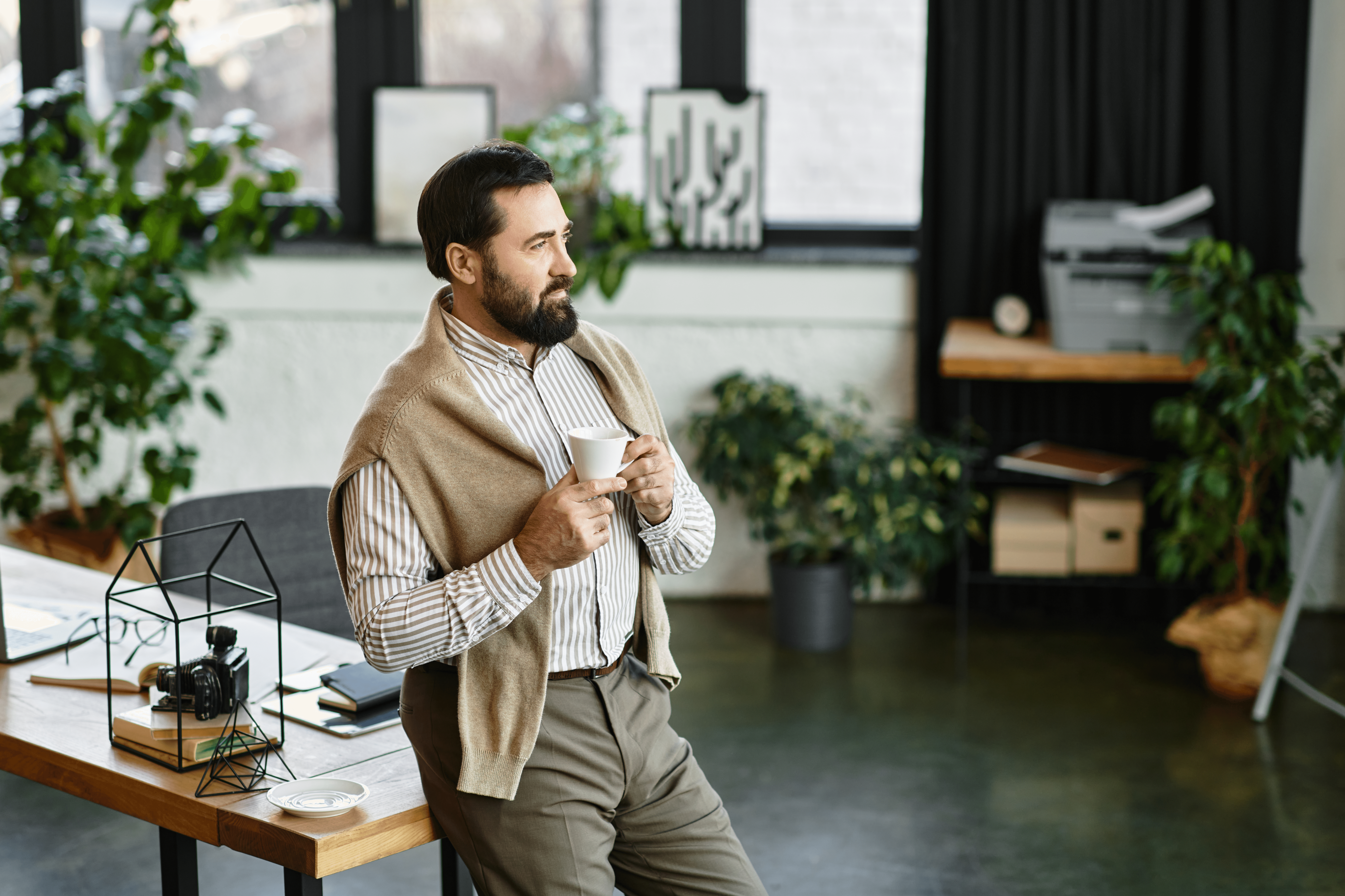 An image of a man leaning against his desk, drinking a cup of coffee.